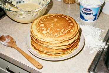 Pile de pancakes au yaourt dorés avec des bulles, entourés d'un bol à pâte, d'une cuillère en bois et d'un pot de yaourt sur un plan de travail de cuisine