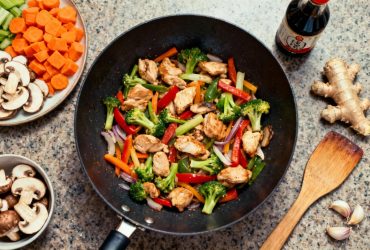 Wok avec poulet et légumes colorés en train de cuire, sur un plan de travail avec ingrédients préparés autour
