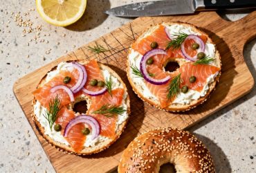 Bagel ouvert garni de saumon fumé, crème fraîche, oignon rouge, câpres et aneth frais posé sur une planche à découper