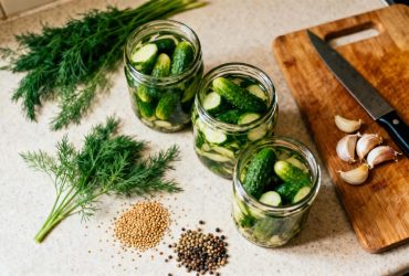 Pots de verre remplis de cornichons maison entourés d'ingrédients frais comme l'aneth, les graines de moutarde et l'ail sur un plan de travail de cuisine