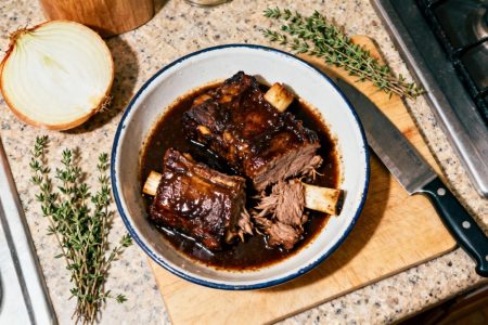 Plat de côte de bœuf braisée tendre avec sauce riche et brillante, accompagnée de thym frais et oignon sur planche à découper