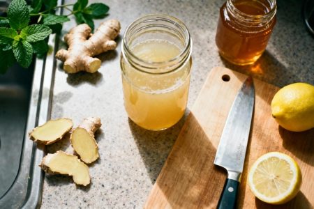 Jus de gingembre frais dans un bocal en verre entouré de morceaux de gingembre pelé, citron et miel sur un plan de travail