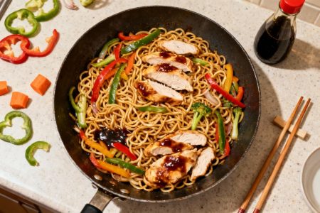 Nouilles chinoises au poulet et légumes colorés dans un wok, avec sauce soja et ustensiles de cuisine autour