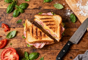 Panini jambon fromage grillé posé sur un plan de travail en bois avec couteau de cuisine et ingrédients autour
