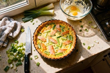 Quiche au saumon et poireaux dorée sur plan de travail avec ingrédients et ustensiles de cuisine dispersés